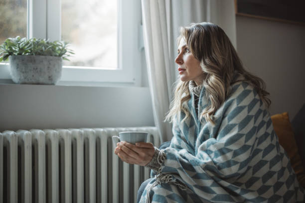 A woman wrapped in a blanket by a bright window, holding a warm cup and looking ahead